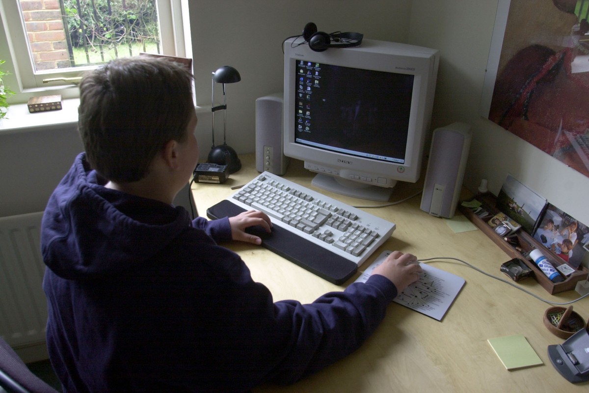 A teenager in a hoodie sits at a desk interacting with a boxy desktop computer, wired mouse, and keyboard.