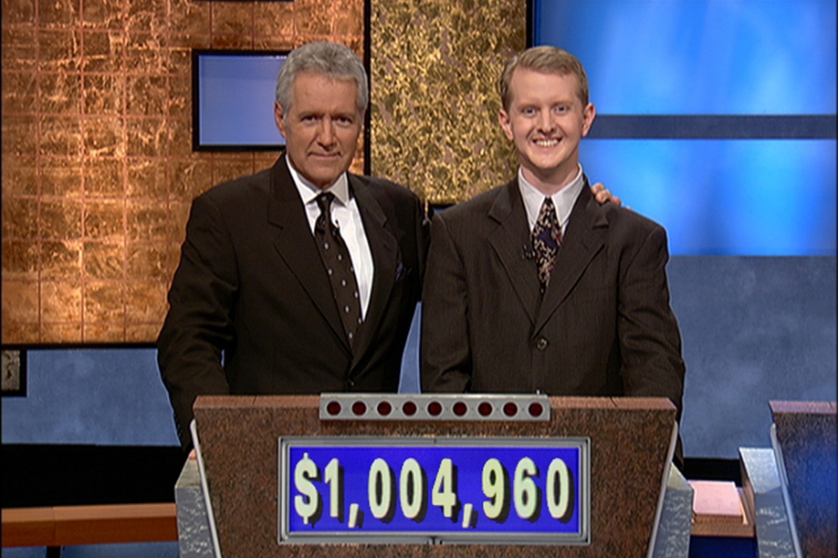 Host Alec Trebek and contestant Ken Jennings stand side by side behind the Jeopardy game show podium showing a score of $1,004,960.